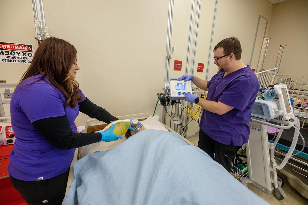Two students stand next to a hospital bed, working on simulated patient.
